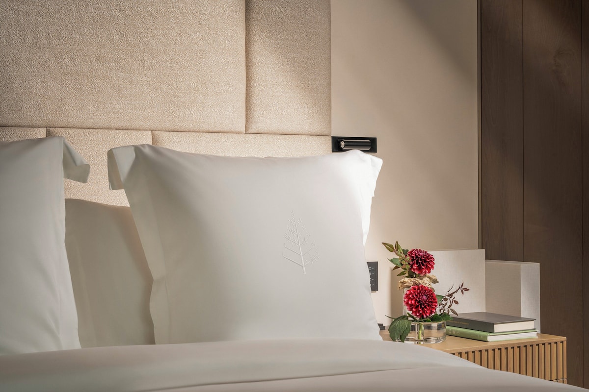 Een net opgemaakt bed met witte lakens en een hoofdbord, een nachtkastje met een boek en een bloemenvaas, en een houten paneelwand.