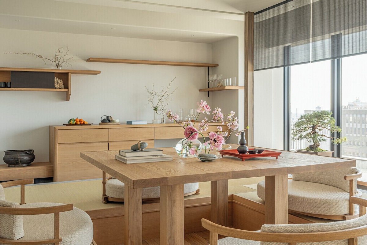 Een strak ingerichte kamer in Japanse stijl, met een houten eettafel, zachte stoelen, een lage houten kast en een bonsaiboom bij het raam.