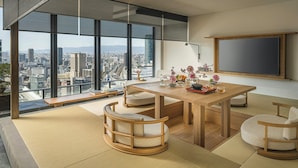 A modern living room with a wooden dining table, cushioned chairs, and a large window offering a city view. - Four Seasons Hotel Osaka (Osaka)
