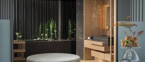 A modern interior with a round wooden bench, a glass table with a vase, and a dark wall with vertical lines.