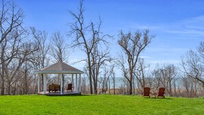 Un kiosque agrémenté de deux fauteuils Adirondack, situé dans un champ herbeux, entouré d’arbres sans feuilles et offrant une vue sur l’eau.