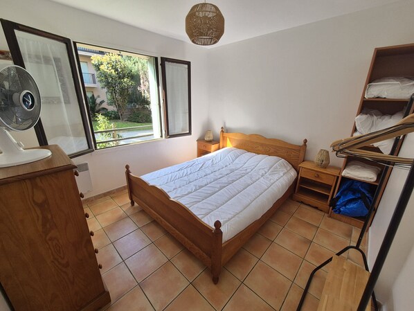 A bedroom with a wooden bed, a nightstand with a lamp, a fan, a window with curtains, and a shelf with folded clothes.