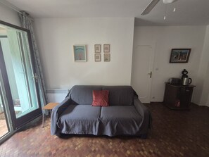 A living room with a grey sofa, a small table, and a cabinet with a kettle. There are framed pictures on the wall and a door to the right.