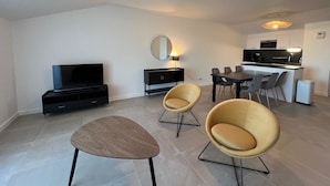 A modern living room with a flat-screen TV, a dining table with chairs, and a kitchen area in the background.