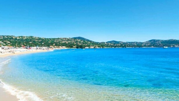 Ein Strand mit klarem, blauem Wasser, sandigem Ufer und einer hügeligen Landschaft im Hintergrund.