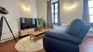 En stue med sofa, fjernsyn på et træstel, en plante og et vindue med gardiner.