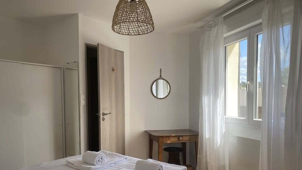 A neatly made bed with white linens and towels, a small wooden table, a mirror on the wall, and a window with sheer curtains.