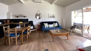 A spacious living area with a dining table, blue sofa, and a glass coffee table. A kitchen area is visible in the background.