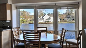 Un espace repas comprenant une table et des chaises, de grandes fenêtres donnant sur un lac et des maisons, ainsi qu’un ventilateur de plafond.
