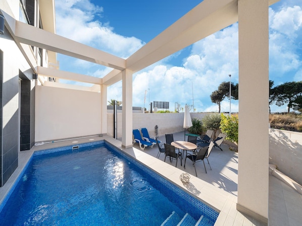 A swimming pool with a tiled deck, a table with chairs, and a view of the sky and distant landscape.