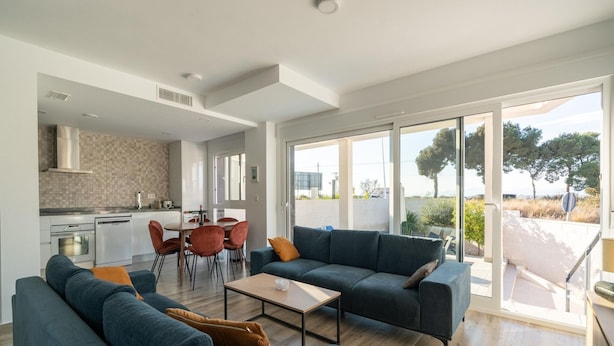 Ein modernes Wohnzimmer mit Sofa, Couchtisch und Essbereich. Im Hintergrund befindet sich eine Küche mit weißen Schränken und einer gefliesten Spritzschutzwand. Große Fenster bieten Blick auf Bäume und ein Gebäude.