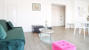 Un saló modern amb un sofà verd, un otomà rosa, una taula de vidre i una cadira blanca.