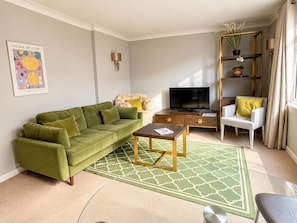 Een woonkamer met een groene bank, een bijzettafeltje, een televisie, een boekenkast en een raam met gordijnen.