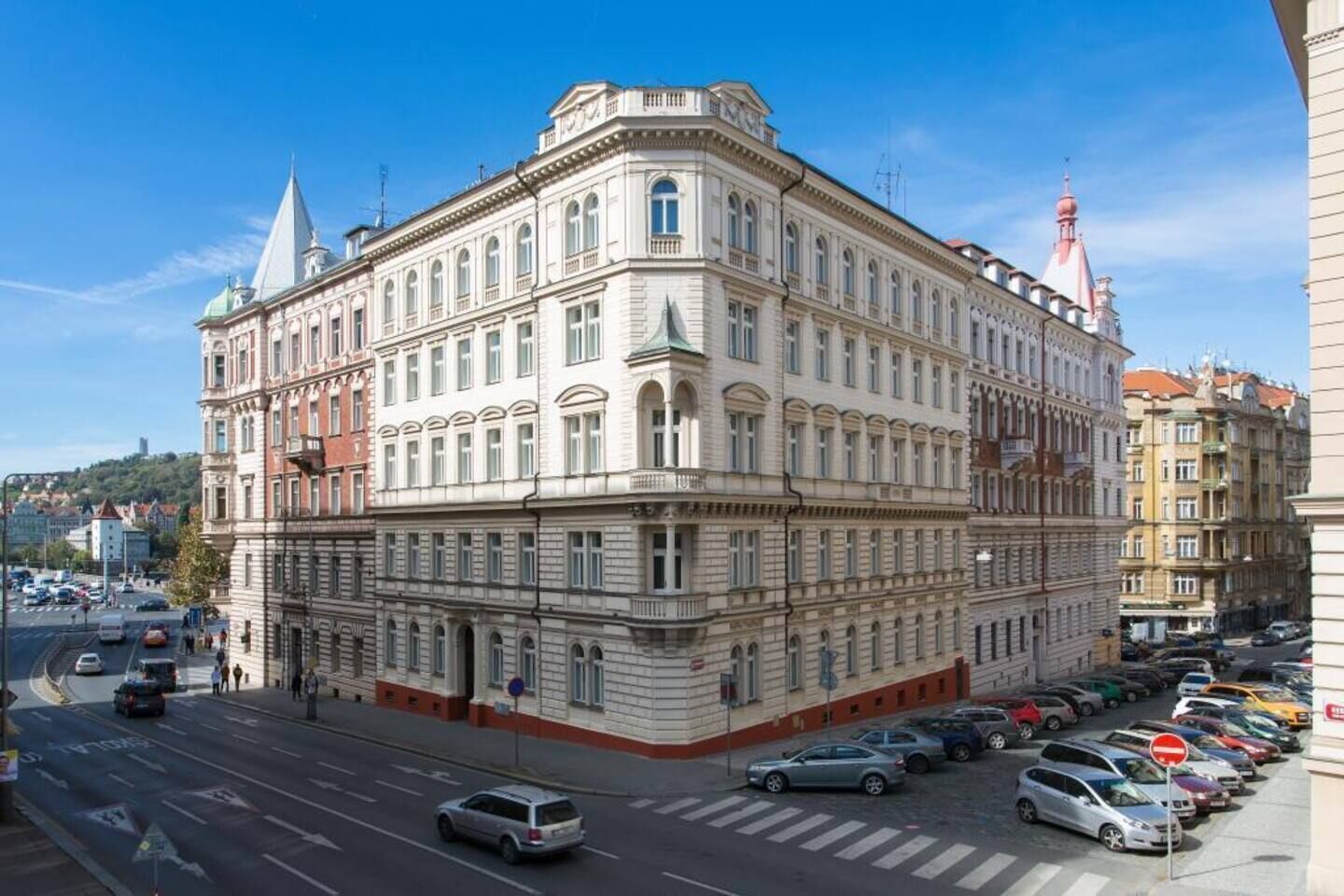 Un immeuble à plusieurs étages, de style architectural classique, qui se distingue par son entrée d’angle imposante et une rangée de voitures garées le long de la rue.