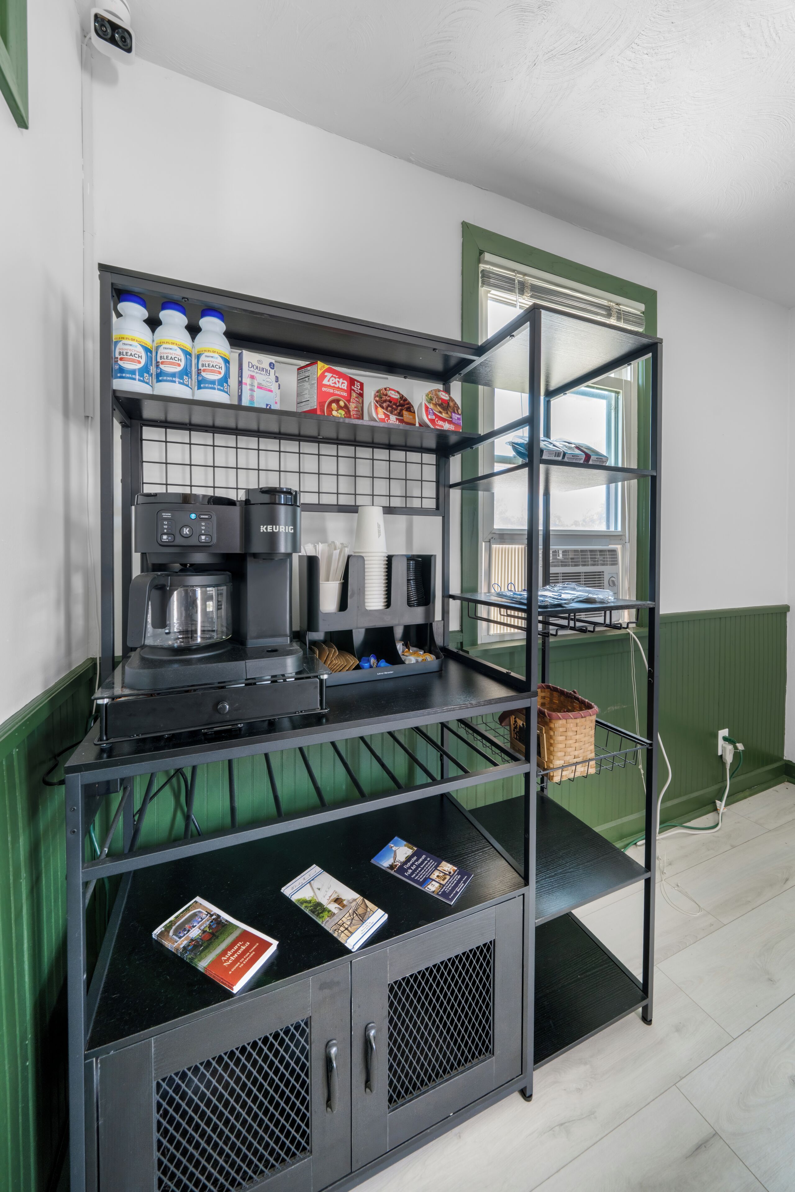 A kitchenette with a coffee machine, shelves with cleaning supplies, and a window with blinds.