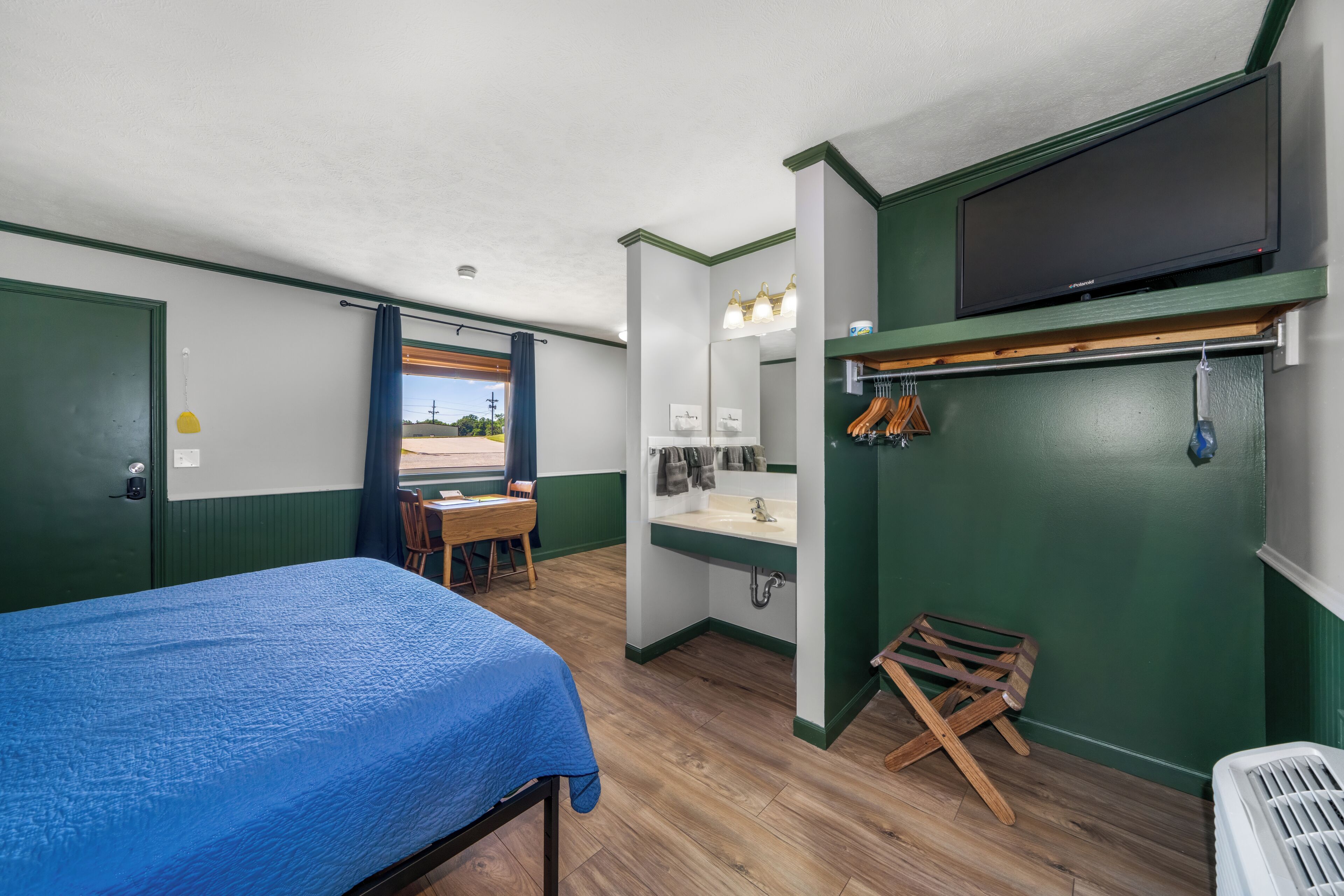 A room with a bed, a desk, a chair, a bathroom with a sink and mirror, and a television mounted on the wall.