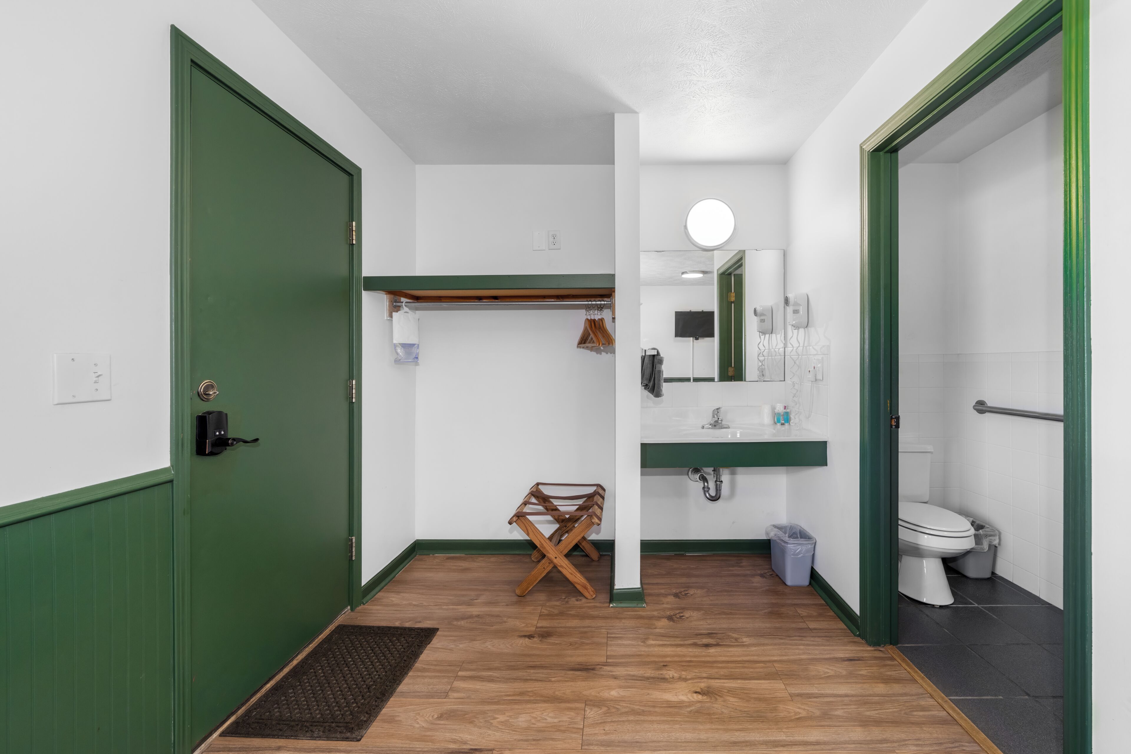 Un couloir étroit avec une porte verte, un banc en bois, un petit miroir et un lavabo blanc surmonté d’un autre miroir.