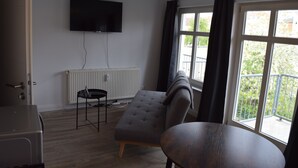 A room with a grey sofa, a wooden table, a black chair, a TV mounted on the wall, and a window with a view of greenery. - Alstadt kiek mal wedder in  (Wismar)