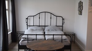 A bed with a metal frame, a round wooden table, and a small stool in a room with a window and a wall-mounted picture frame. - Alstadt kiek mal wedder in  (Wismar)