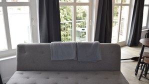 A grey sofa with two grey throw blankets, a dining table with chairs, and a view of greenery through large windows. - Alstadt kiek mal wedder in  (Wismar)