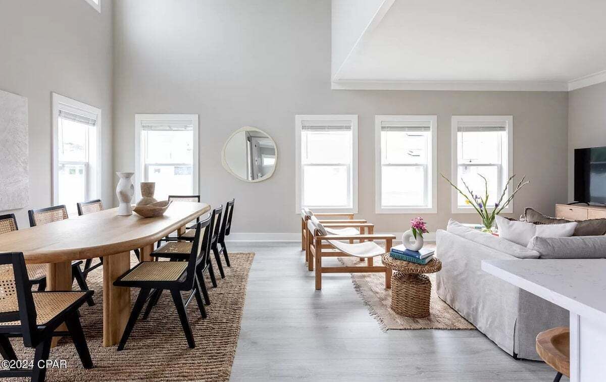 Un salon spacieux avec une grande table à manger, un canapé et un téléviseur. De grandes fenêtres laissent entrer la lumière naturelle.
