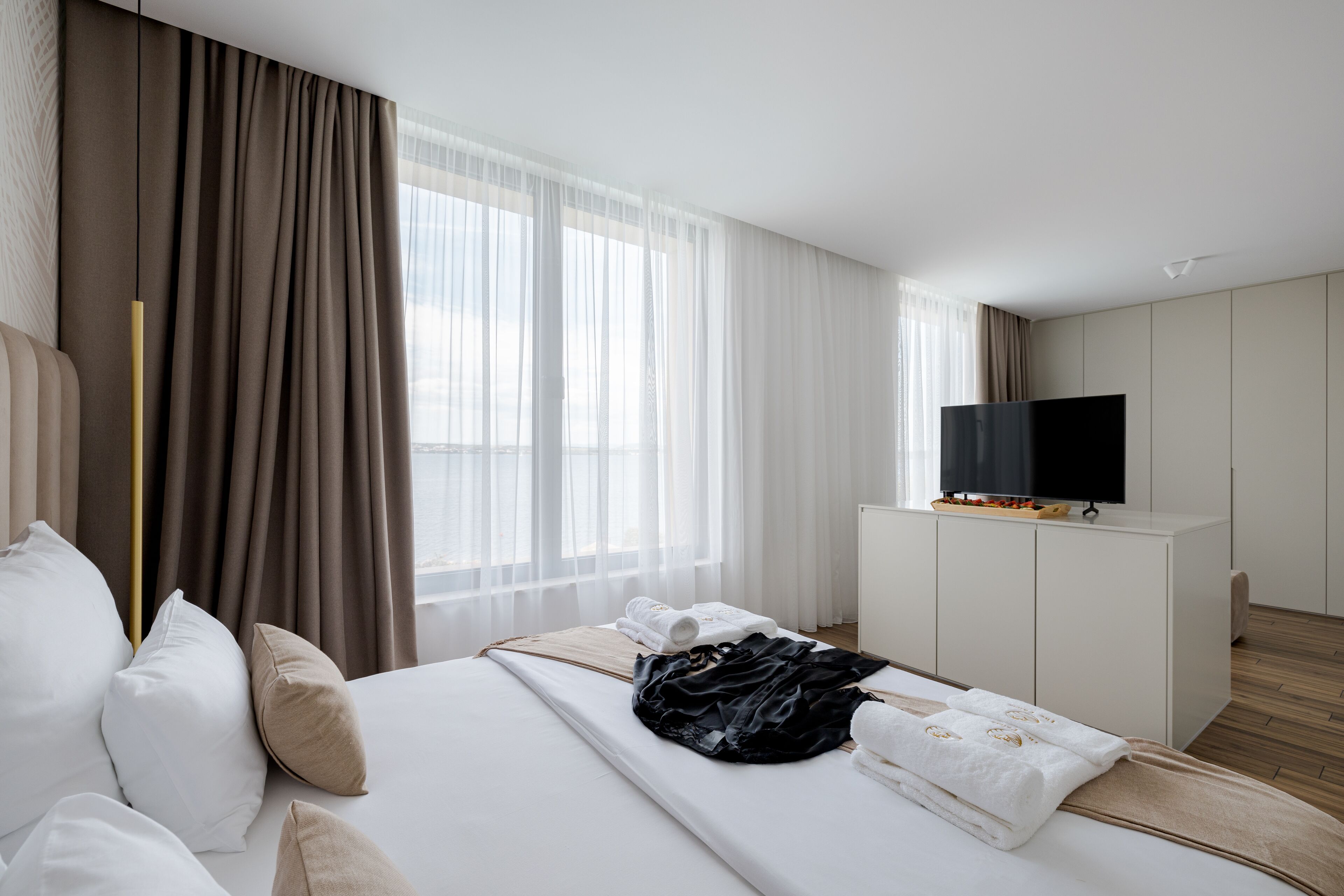 A modern hotel room with a large bed, a television on a cabinet, and a window with sheer curtains.