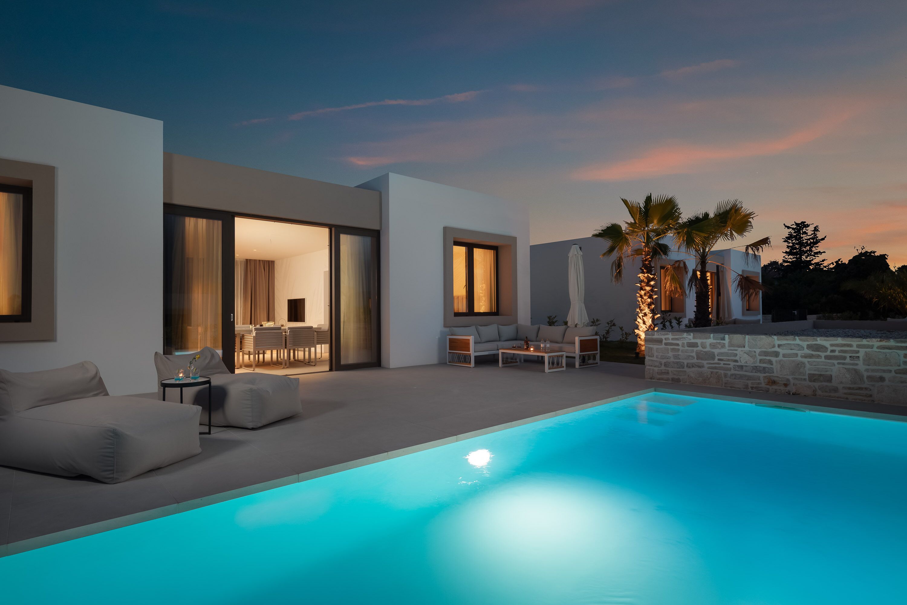 A modern outdoor pool area with a sofa and chairs, a dining area, and a stone wall.
