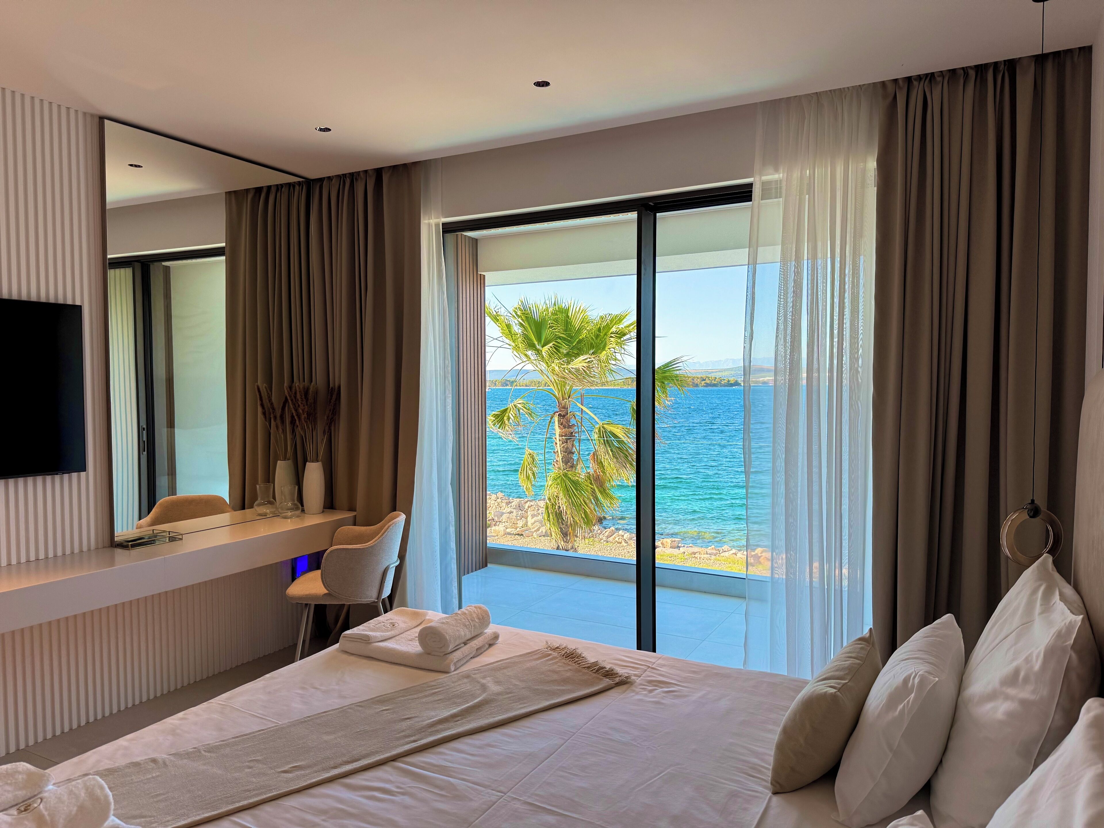 A modern hotel room with a large bed, a desk with a chair, a TV, and a balcony with a view of the sea and palm trees.