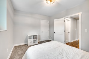 A bedroom with a large bed, white walls, and a carpeted floor. There are two white doors and a white nightstand with a lamp.