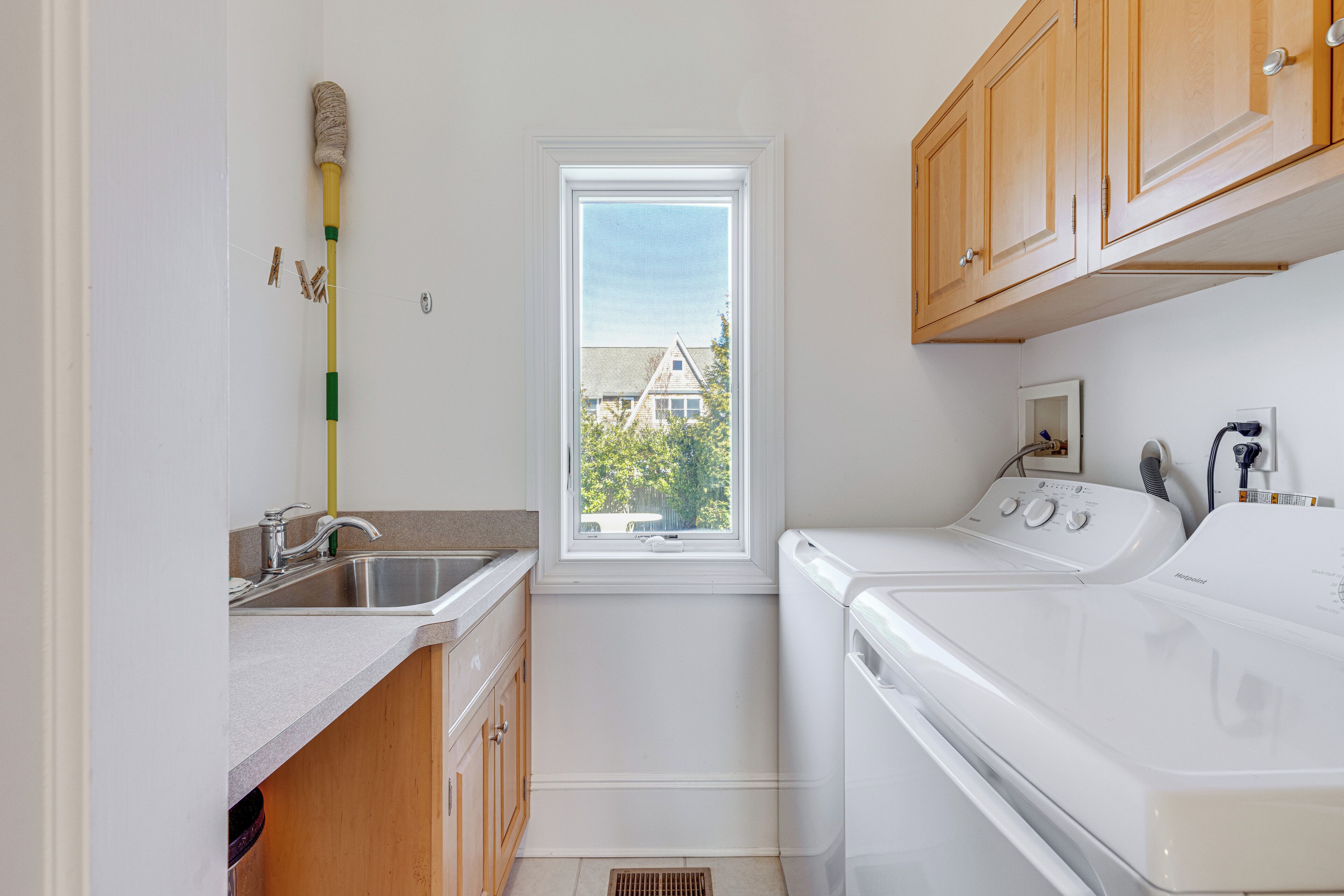 A laundry room with a washing machine, a sink, wooden cabinets, and a window with a view of a house.