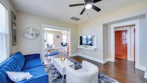 Ein Wohnzimmer mit blauem Sofa, weißem Sessel und Couchtisch. Es gibt einen Deckenventilator, einen an der Wand montierten Fernseher und eine Holztür.