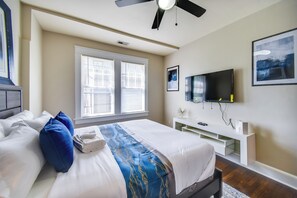 A bedroom with a bed, pillows, a TV, a ceiling fan, and a window with blinds.
