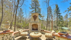 An outdoor patio with stone walls, a stone fireplace, and two wicker chairs. - Lake Arrowhead Home: Mountain View, Walk to Water! (Lake Arrowhead)
