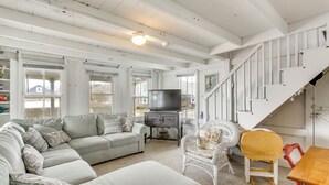 A spacious living room with a large grey sofa, a white armchair, and a wooden table. A staircase with white railings leads to an upper level. - Charming Westbrook Cottage, Steps to Private Beach (Westbrook)