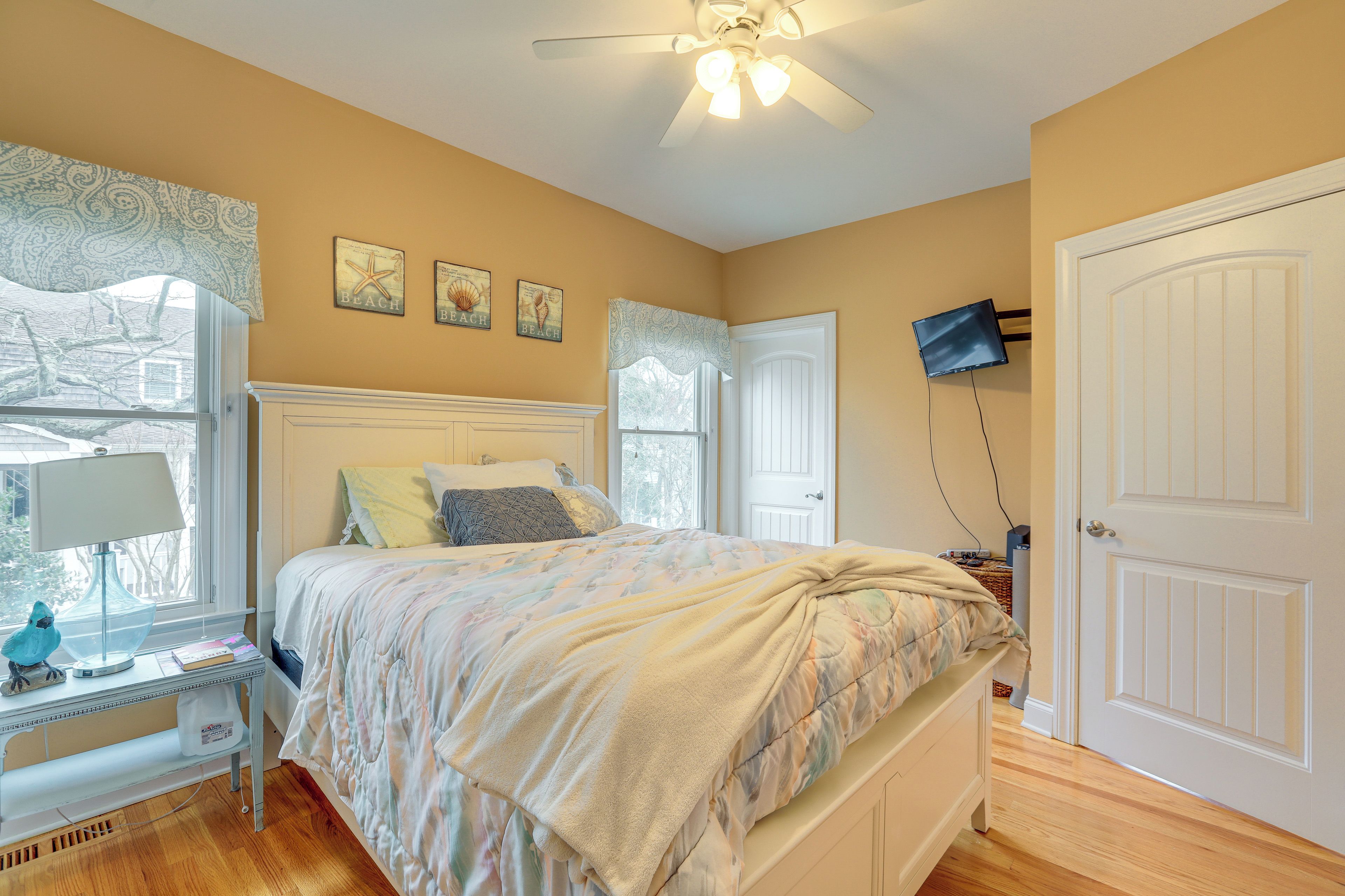 A bedroom with a bed, a desk with a lamp, a ceiling fan, and a TV mounted on the wall.