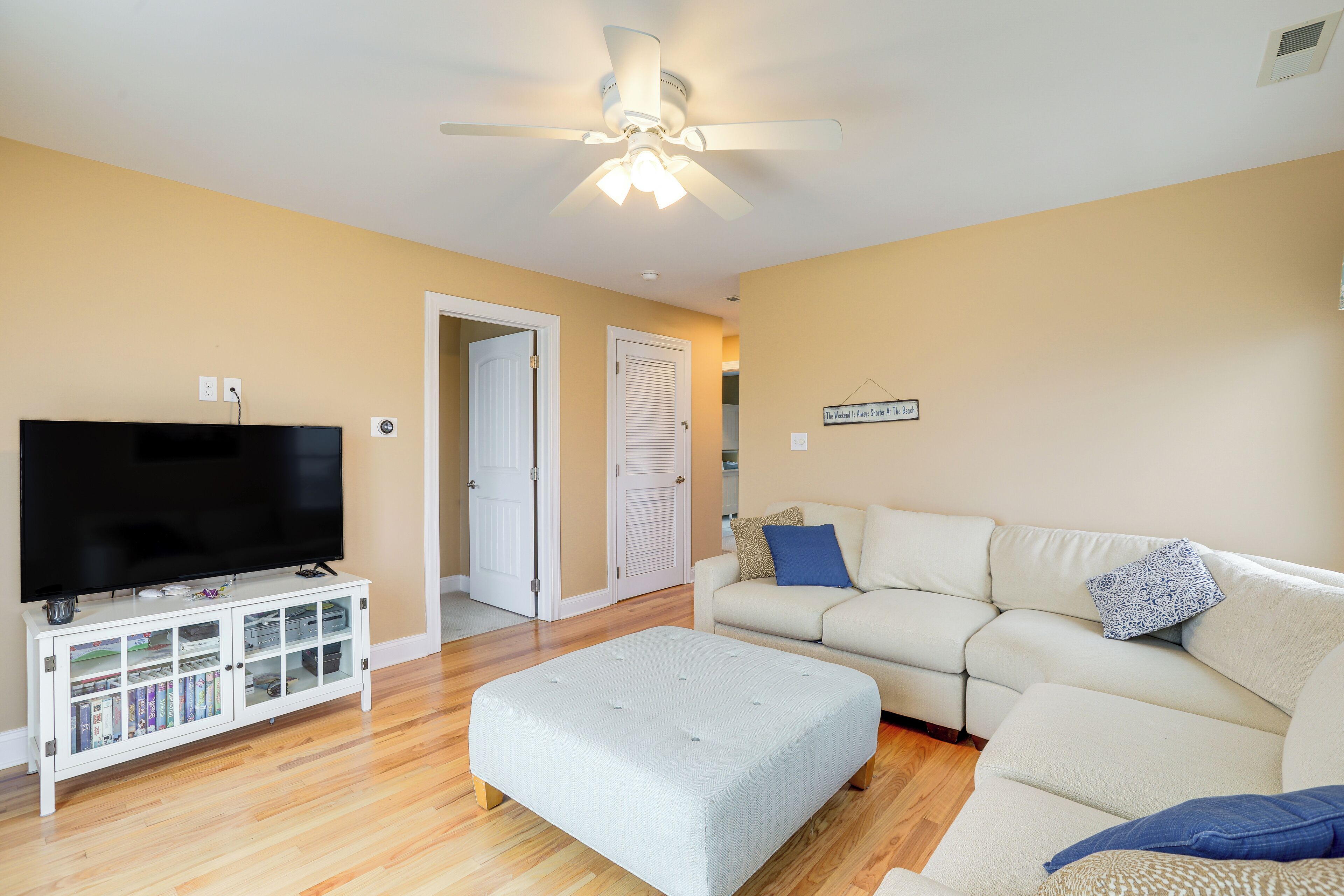 A living room with a flat-screen TV on a white entertainment unit, a beige sofa with a blue pillow, and a ceiling fan.