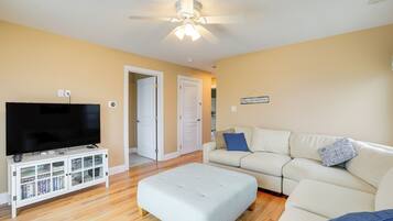 A living room with a flat-screen TV on a white entertainment unit, a beige sofa with a blue pillow, and a ceiling fan.