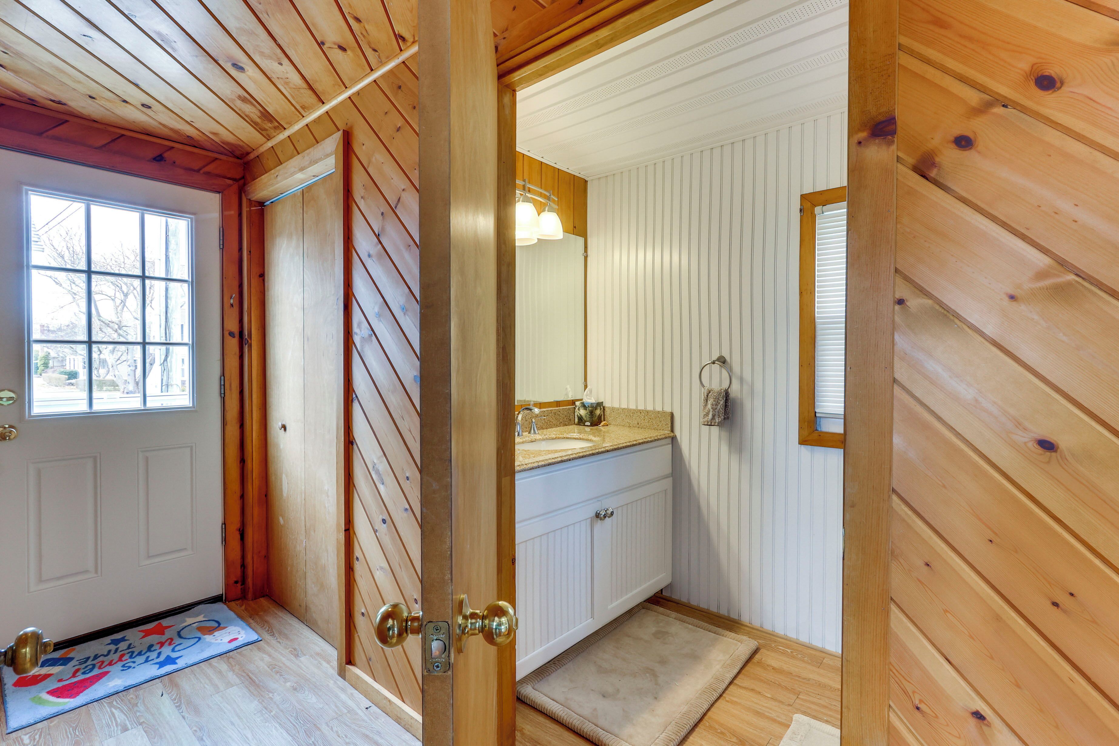 A wooden interior with a white door, a wooden floor, a bathroom with a sink and mirror, and a window with blinds.