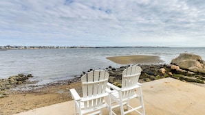 Zwei weiße Adirondack-Stühle, die auf einen Strand mit felsigem Ufer und Blick auf Wasser sowie entfernte Gebäude zeigen.