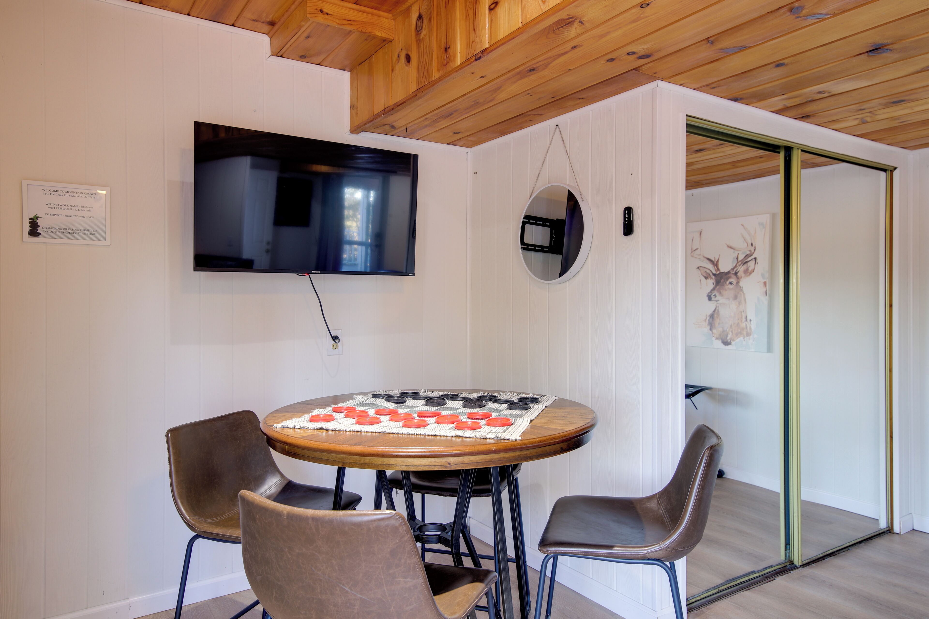 A room with a round wooden table, four chairs, a mounted TV, a wall-mounted mirror, and a door leading to another room.