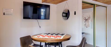 A room with a round wooden table, four chairs, a mounted TV, a wall-mounted mirror, and a door leading to another room.