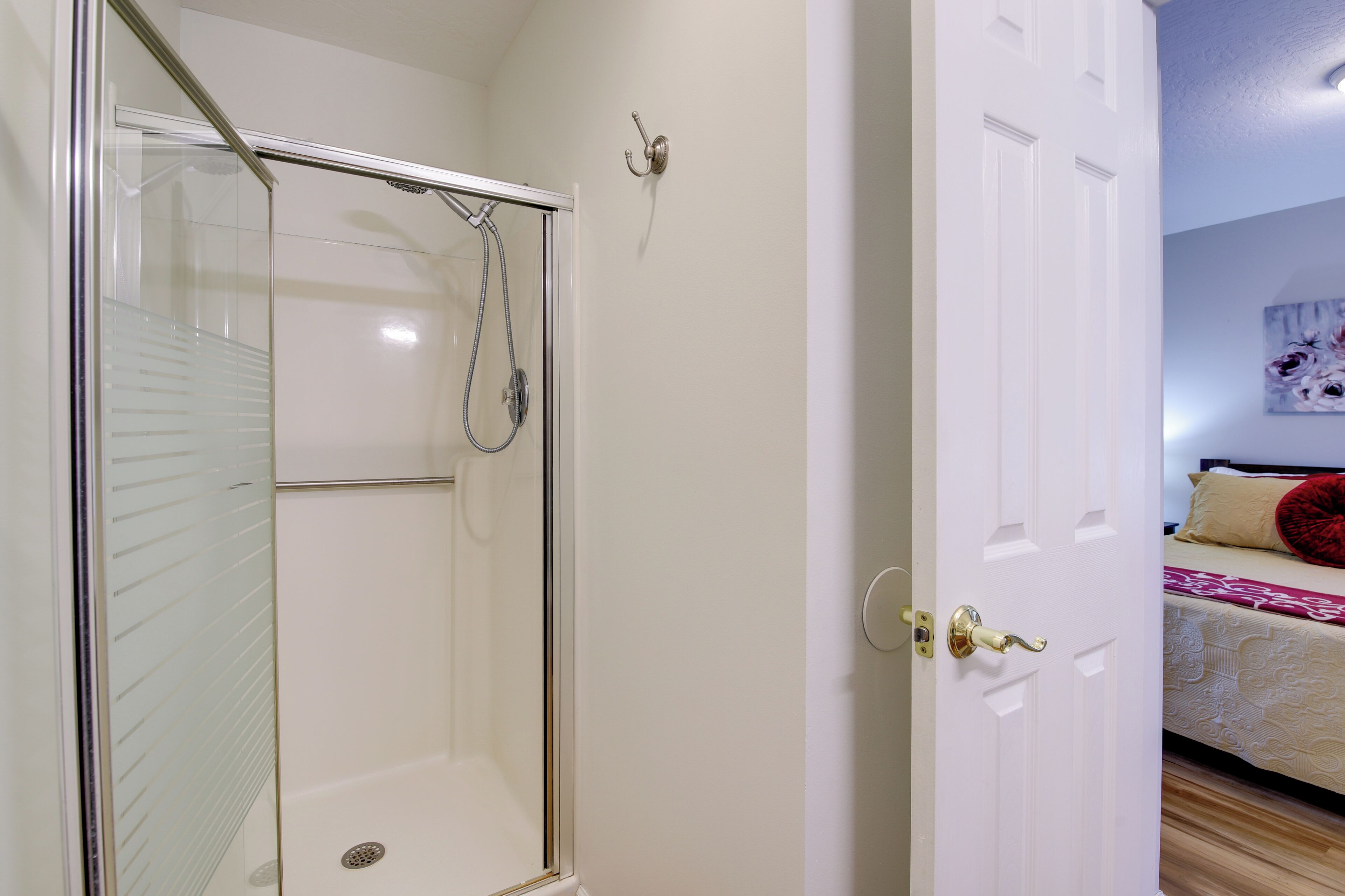 A glass-enclosed shower with a handheld showerhead, a white door with a brass handle, and a bed with a red pillow in the adjacent room.