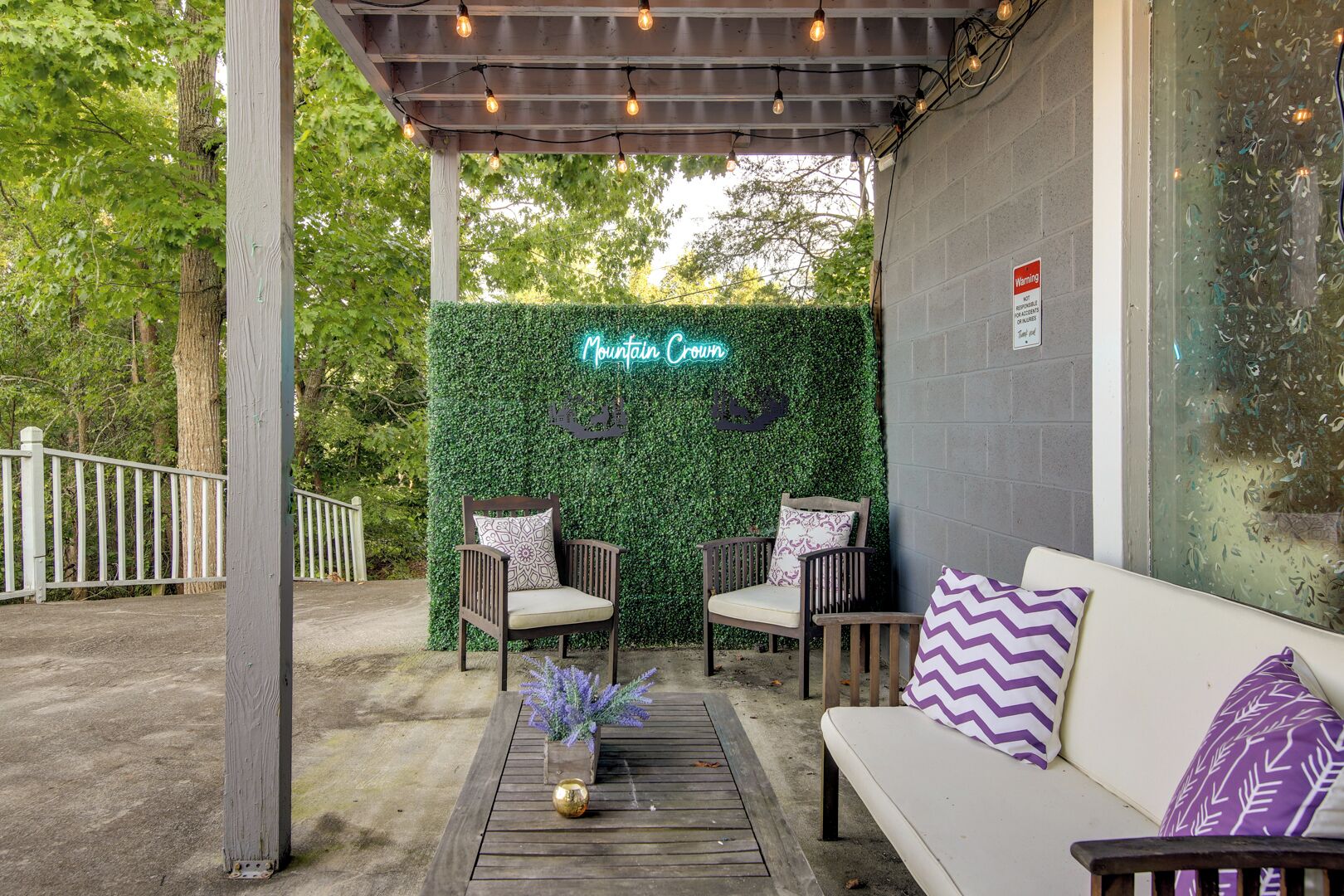 Ein Sitzbereich im Freien mit zwei Holzstühlen und einem weißen Sofa, einer Neonreklame mit der Aufschrift „Mountain Crown“ und einer dekorativen Hecke.