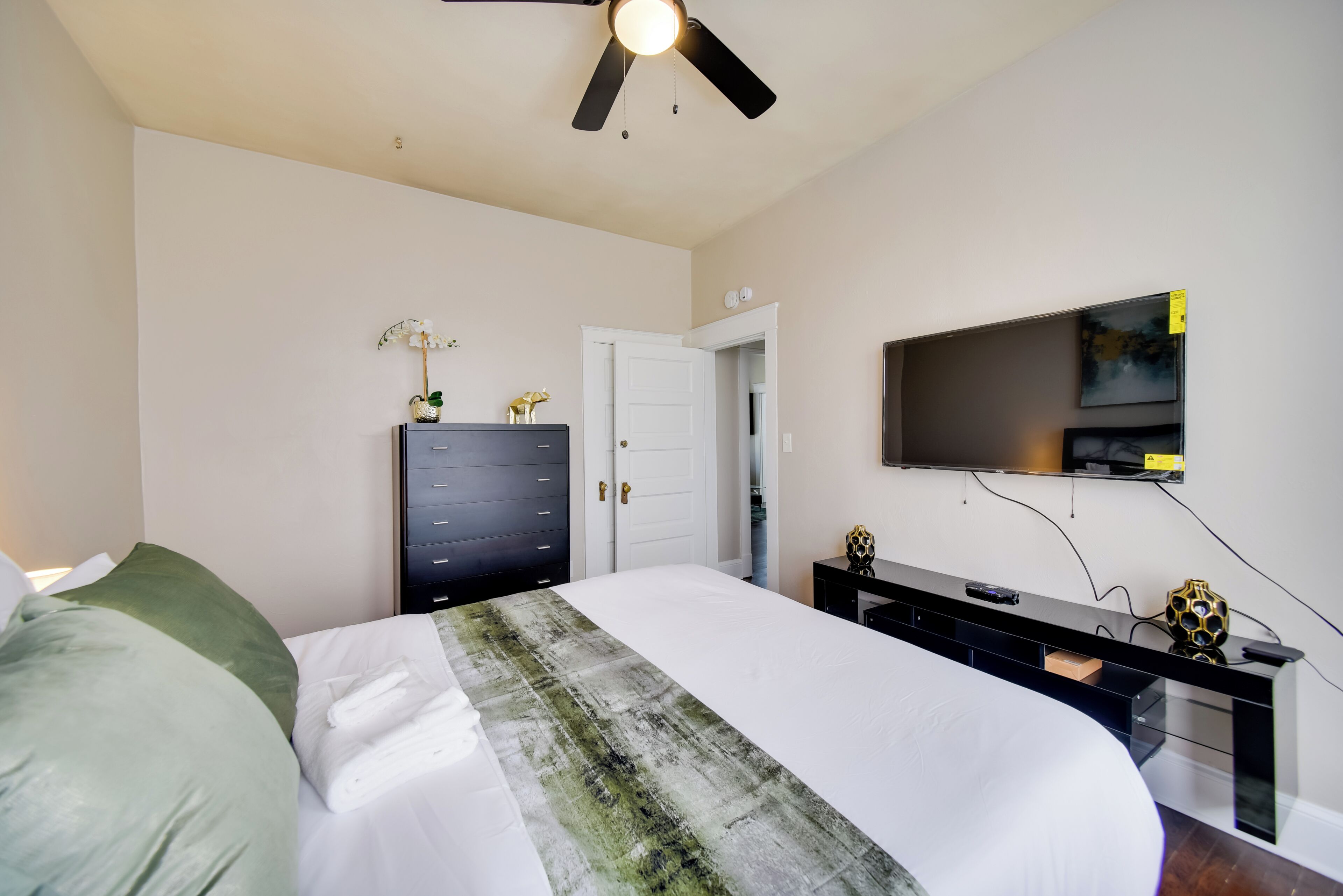 A bedroom with a bed, a dresser, a TV mounted on the wall, and a ceiling fan.