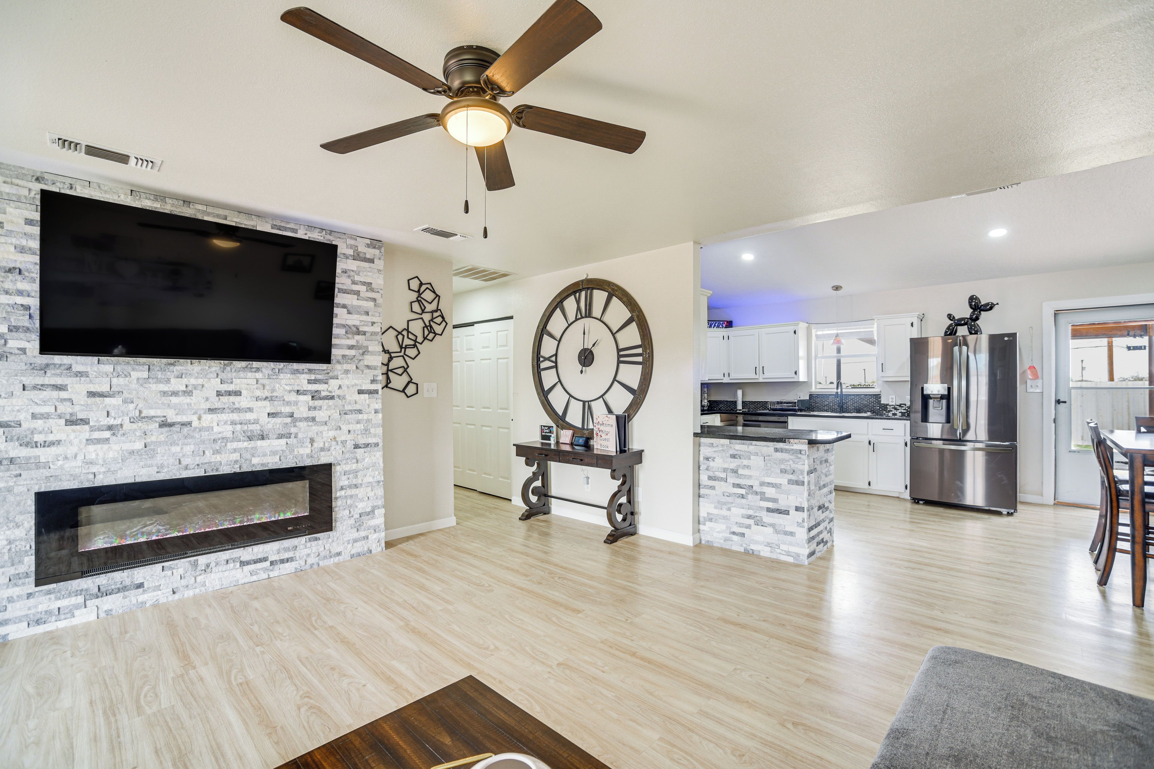 A modern living room with a flat-screen TV, a fireplace, a large wall clock, and a kitchen area with stainless steel appliances.