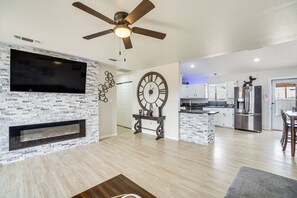 Een moderne woonkamer met een flatscreen televisie, een open haard, een grote wandklok en een keuken met roestvrijstalen apparatuur.