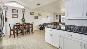 Une cuisine spacieuse avec des meubles blancs, un coin repas avec des meubles en bois et un salon doté d’un canapé gris.