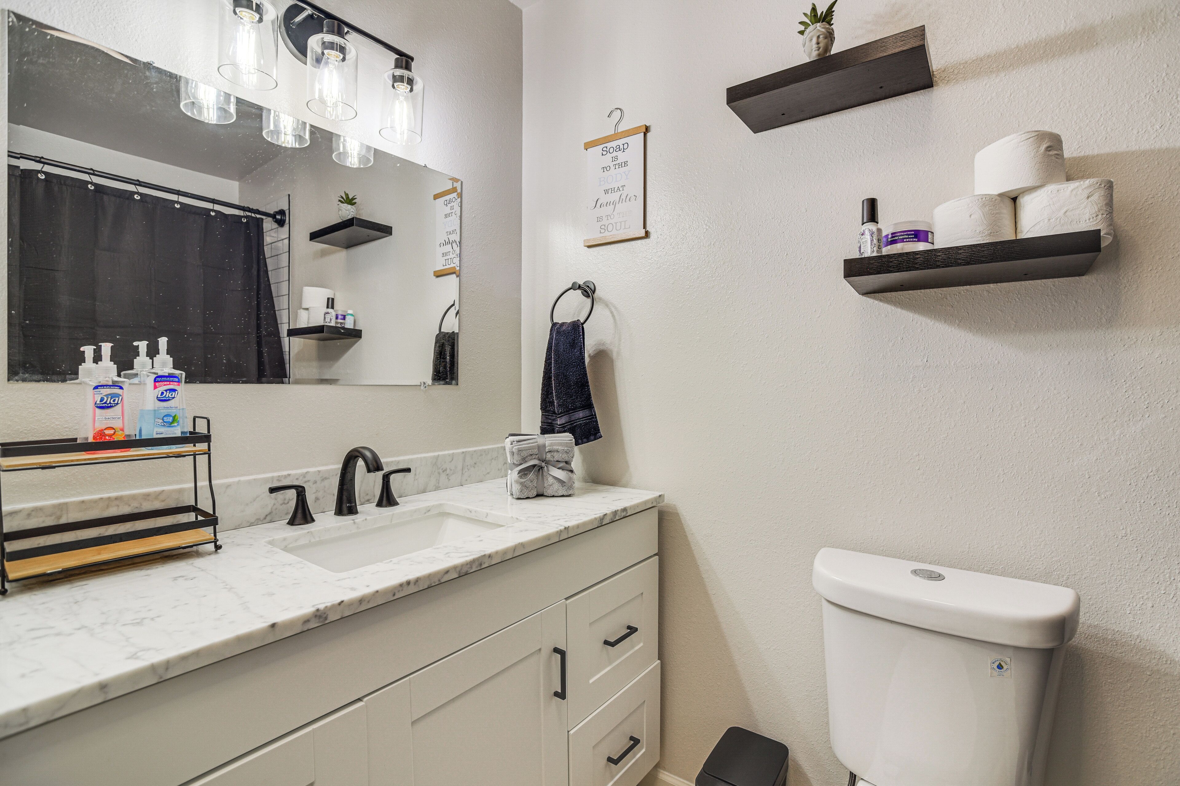 Un baño con lavamanos blanco, grifo negro y retrete blanco. Hay estantes con artículos de aseo personal y una toalla colgada en la pared.