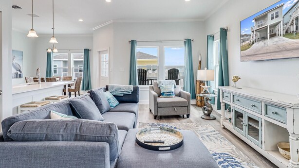 A spacious living room with a gray sectional sofa, a small gray armchair, and a coffee table with a decorative tray. There is a white console table with drawers and a lamp. The room has a large window with teal curtains, a ceiling fan, and a painting of a house on the wall. - Pelican Reef Or104 3 Bedroom Home by RedAwning (Rockport)