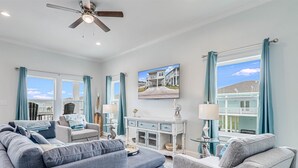 A spacious living room with a large gray sofa, a smaller gray armchair, and a white console table with decorative items. A ceiling fan and a large window with teal curtains are visible. - Pelican Reef Or104 3 Bedroom Home by RedAwning (Rockport)